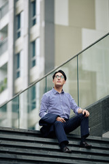 Dreamy Asian entrepreneur taking off jacket and resting on steps and looking up