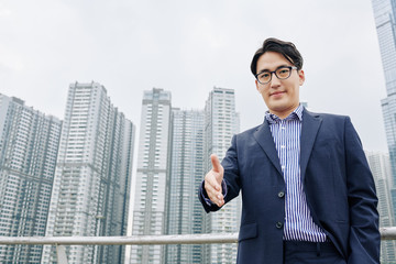 Fototapeta na wymiar Smiling Asian businessman outstretching hand for handshake, skyscrapers in background