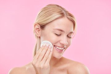 Pretty young smiling female with cotton pad enjoying procedure of cleaning face