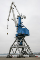Fototapeta na wymiar A large high port crane on rails against a gray sky. Powerful system for lifting ship cargo. The crane is white with blue. Vertical frame.
