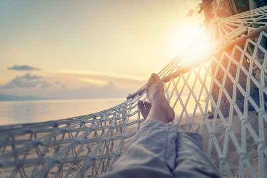 Female legs in a hammock on a background of the sea, palm trees and sunset. Vacation concept, point of view