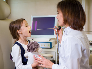 Little girl at the reception in the ENT clinic. Female doctor shines in the throat with a special device. Theme of children's medicine - Powered by Adobe