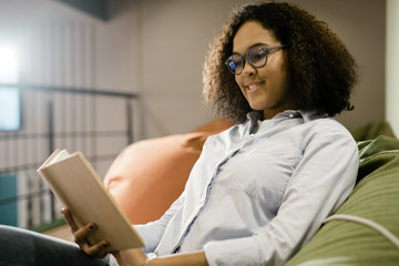 Happy young clever female student in new eyeglasses reading book on couch