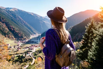Foto op Canvas Stijlvolle trendy hipster vrouw reiziger in een vilten hoed met bruine rugzak staat op de achtergrond van de bergen en het uzungol-meer in Trabzon tijdens reizen naar Turkije © Goffkein