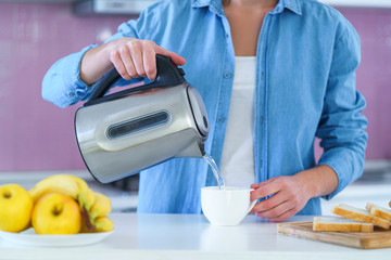 Woman pouring boiled water in a cup from an electric kettle for brewing hot tea at home at kitchen