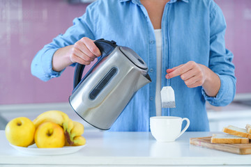 Woman putting tea bag in a cup and using an electric kettle for brewing hot tea at home at kitchen
