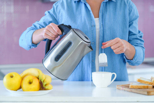 Woman putting tea bag in a cup and using an electric kettle for brewing hot tea at home at kitchen