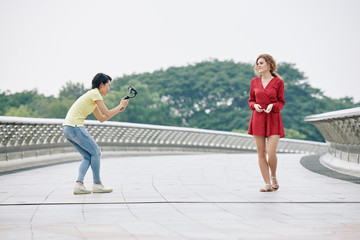 Fototapeta premium Young Vietnamse woman with smartphone filming pretty blogger talking when walking along the bridge