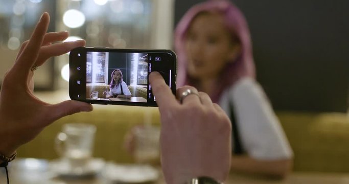 Young couple in a modern caffe, man photographs a girl with pink hair on the smartphone, teenagers have a rest in the city.