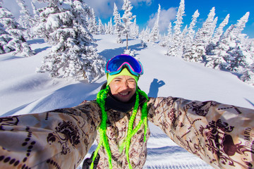 Selfie portrait of young cheerful woman with green braids on a snowy slope at winter ski resort.
