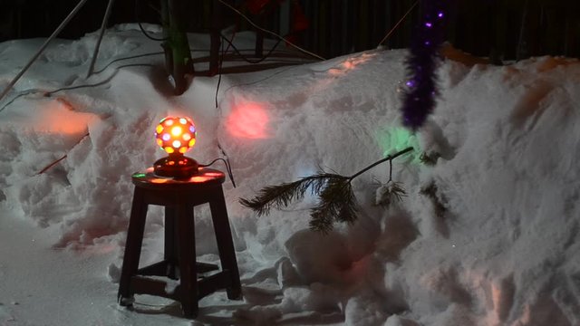 Disco ball spinning on a wooden chair under the Christmas tree throwing multicolored glare on the snow holiday night.