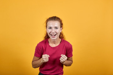 Pretty young lady in red shirt doing winner gesture, smiling, keeping fists up isolated on orange background in studio. People sincere emotions, health concept.