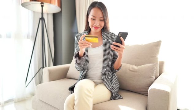A well dressed young Asian woman holds her credit card in one hand while looking at her smartphone in the other.