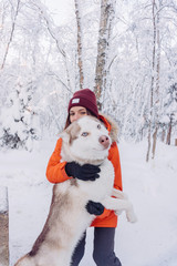 girl in a bright orange jacket sits and plays and hugs with a husky breed dog in the winter forest after snowfall during frost