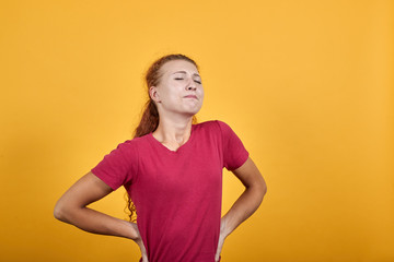 Pretty young lady in red shirt stretch her back, looking painful isolated on orange background in studio. People sincere emotions, health concept.