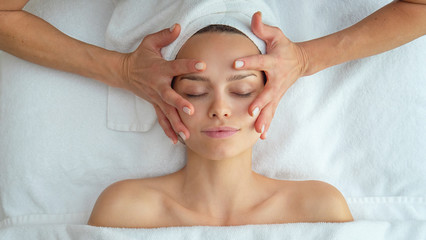 Close up portrait of a beautiful young smiling woman with a towel on her head is receiving a facial massage and spa treatment for perfect skin in a luxury wellness center.