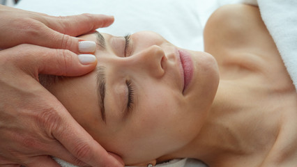 Close up portrait of a beautiful young smiling woman with a towel on her head is receiving a facial massage and spa treatment for perfect skin in a luxury wellness center.