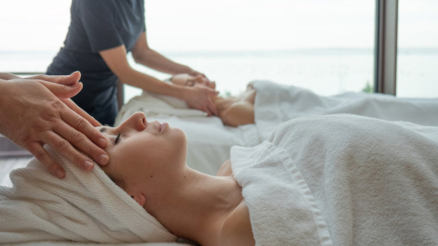 Portrait of two young girlfriends with a towel on head are receiving a facial massage and spa treatment for perfect skin in a luxury wellness center.