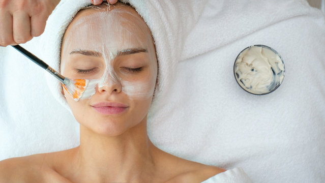 Close up of cosmetologist applies facial clay mask on an young beautiful woman face before ultrasonic cleaning procedure for skin pores and deep moisturizing. - Powered by Adobe