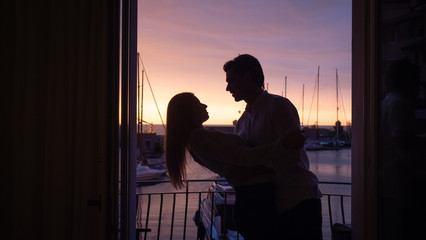 Close up of an young couple is enjoying time together on a terrace of a hotel room during their romantic vacation with panoramic view on a sea with yachts at sunset.