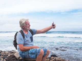 Happy senior man sitting on the beach with cellphone taking a selfie. Large waves of the sea in background. Cloudy sky