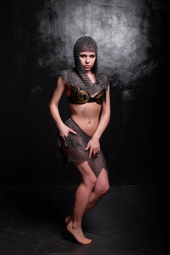 Beautiful girl in a black leather bra and a chainmail skirt and a chainmail hood posing while standing in a dark room. Studio photo, retouching, model with clean skin.