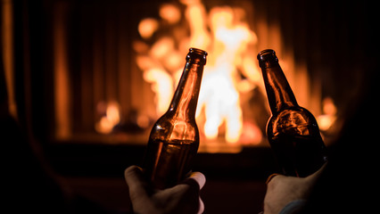 Two bottles of beer in the hands of men on a background of fire in a fireplace