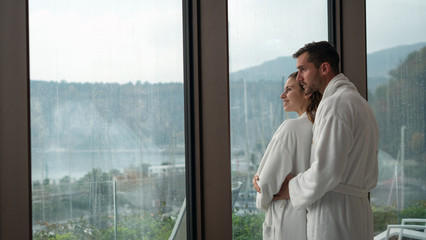 An young happy embraced couple in white bathrobes after various treatments are enjoying the panoramic view through a window in a luxury wellness center