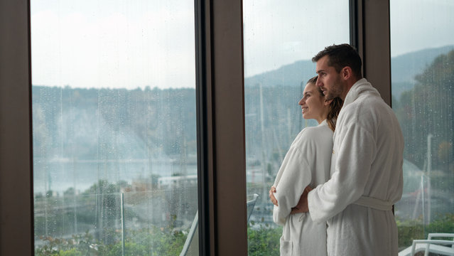 An young happy embraced couple in white bathrobes after various treatments are enjoying the panoramic view through a window in a luxury wellness center
