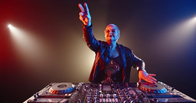 Funny caucasian grandma in authentic outfit and makeup working as a cool dj in a nightclub, dancing and cheering at turntables 4k footage