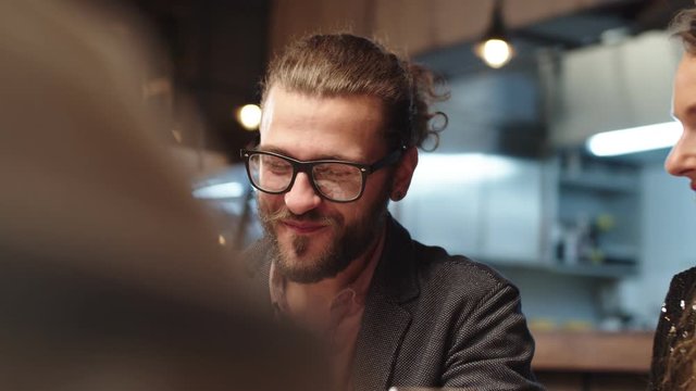 Handsome caucasian bearded hipster man with eyeglasses drinking wine while chatting and sitting with his friends in restaurant.