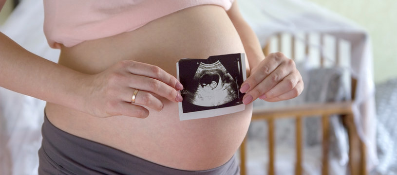 Big pregnant tummy and woman's hands holding ultrasound image of healthy unborn baby in the interior of the children's room. Maternity prenatal care and woman pregnancy concept. Panoramic banner.