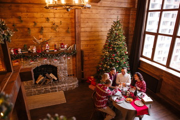 Friends sitting in a large wooden house at a festive table next to a Christmas tree and a fireplace celebrate the new year view from above