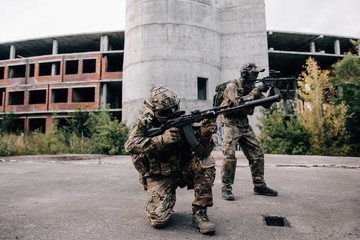 Two military soldiers in camouflage with assault rifles looking through the scope preparing to attack standing in the background of a destroyed building