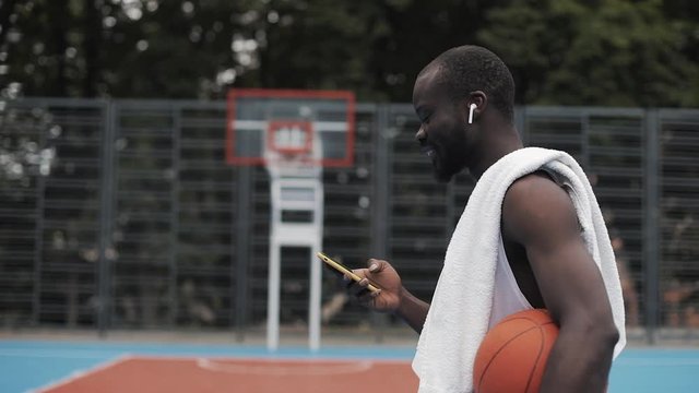 Afro - American Guy in Earphones Holding Ball in one hand and his Smartphone in other Listening his Favourite Music and Smiling, Walking at Basketbal Court. Sport and Technology Concept. Side View.
