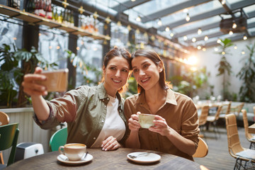 Portrait of two young women smiling at smartphone camera while taking selfie photo in outdoor cafe