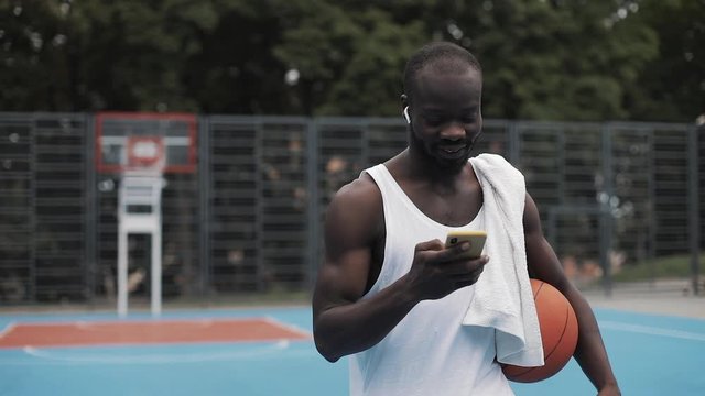Afro - American Guy in Earphones Holding Ball in one hand and his Smartphone in other Listening his Favourite Music Singing and Smiling, Walking at Basketbal Court. Sport and Technology Concept.