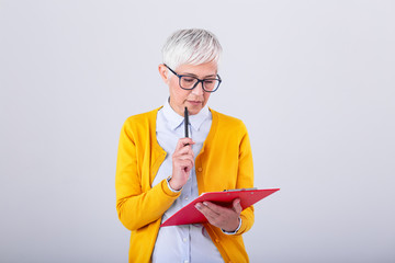 Photo of a thinking mature business woman isolated over grey background holding clipboard and pen....