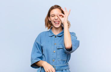 young blonde woman smiling happily with funny face, joking and looking through peephole, spying on...