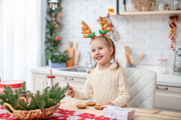 dreamy child in a light sweater and headband with reindeer horns,sitting at the kitchen table smiling spread her hands to the sides.