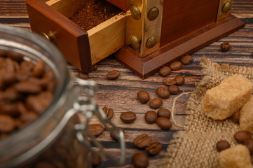 Coffee beans in a glass jar, coffee grinder, pieces of brown sugar on a wooden background. Close up.