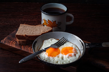 Fried scrambled eggs in vintage pan, with bread, old fork, metal mug with tea, side view, dark background