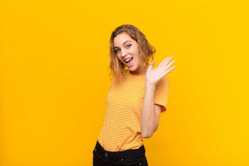 young blonde woman smiling happily and cheerfully, waving hand, welcoming and greeting you, or saying goodbye against flat color wall