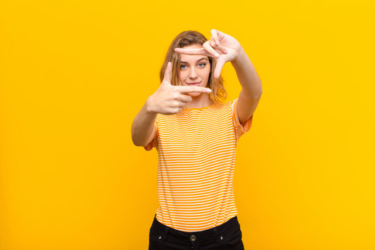 young blonde woman feeling happy, friendly and positive, smiling and making a portrait or photo frame with hands against flat color wall