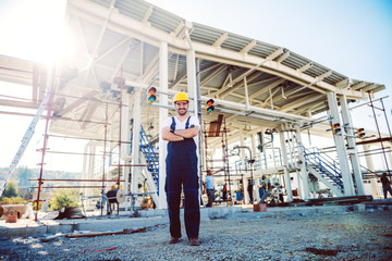 Full length of handsome caucasian worker in overalls and helmet on head standing outdoors with arms crossed. Oil production.