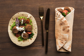top view of falafel with vegetables and sauce wrapped and unwrapped in pita on wooden table with cutlery