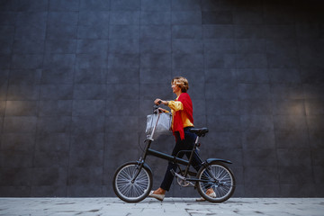 Obraz na płótnie Canvas Full length of gorgeous healthy blonde caucasian senior woman pushing bicycle and passing by gray wall.