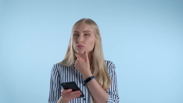 Smart young woman looking at smartphone and start planning something on blue background. She holding her chin with her hand.
