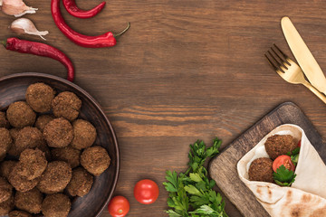 Naklejka na ściany i meble top view of falafel with vegetables and sauce in pita near balls, parsley, tomatoes, chili pepper, garlic and cutlery on wooden table
