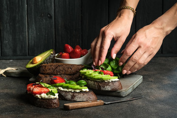 Toast or sandwich with avocado, cheese, strawberries, herbs and seeds on a dark background. Female hands serve a dish. An idea for bruschetta or for a healthy snack. Healthy vegan breakfast.